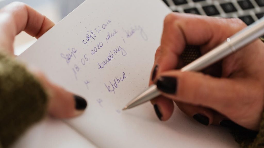 Crop anonymous female with manicured hands writing down notes in notebook with white sheets by using stylish silver pen near modern laptop