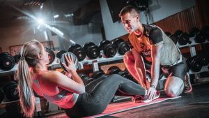 A personal trainer assists a woman doing sit-ups with a medicine ball in a gym setting.